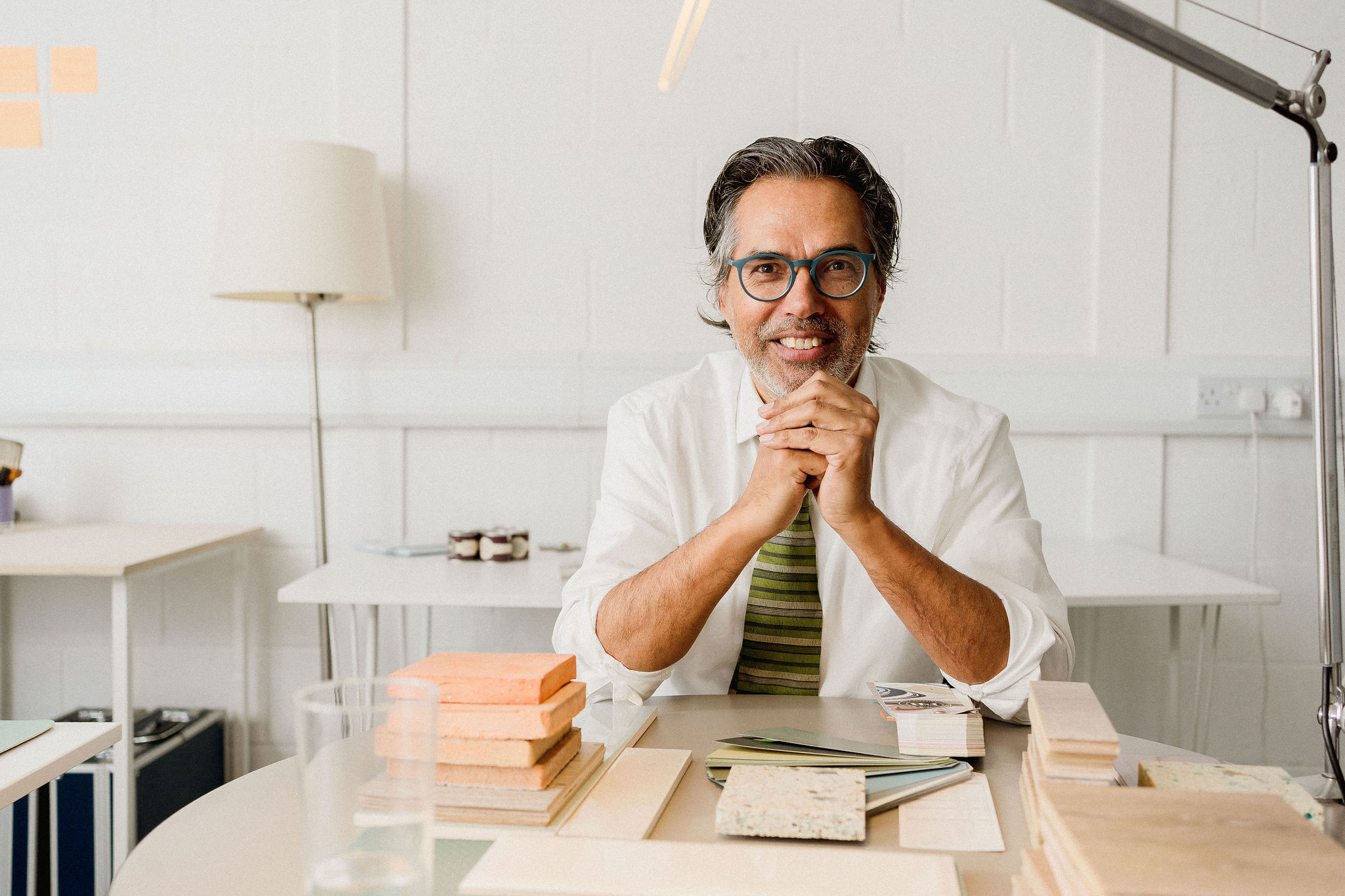 Architect in his office wearing white shirt and a green tie, at his meeting table with paint samples, bricks and tiles examples, looking into the camera smiling